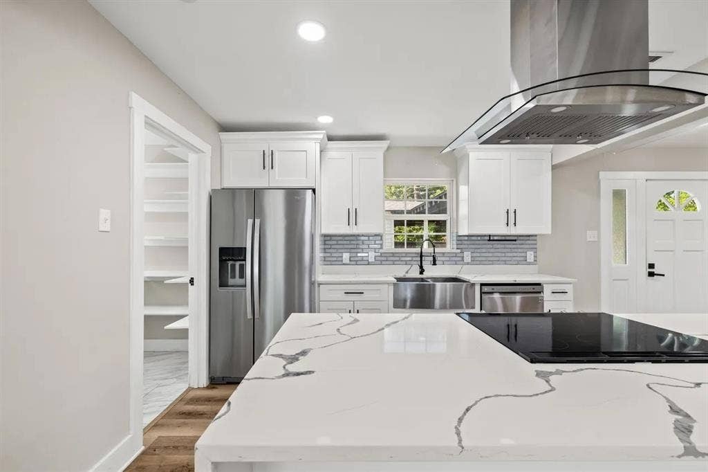 Kitchen featuring an oversized stone-finish waterfall island, stainless steel range hood, farmhouse sink, shaker-style cabinetry, and a walk-in pantry