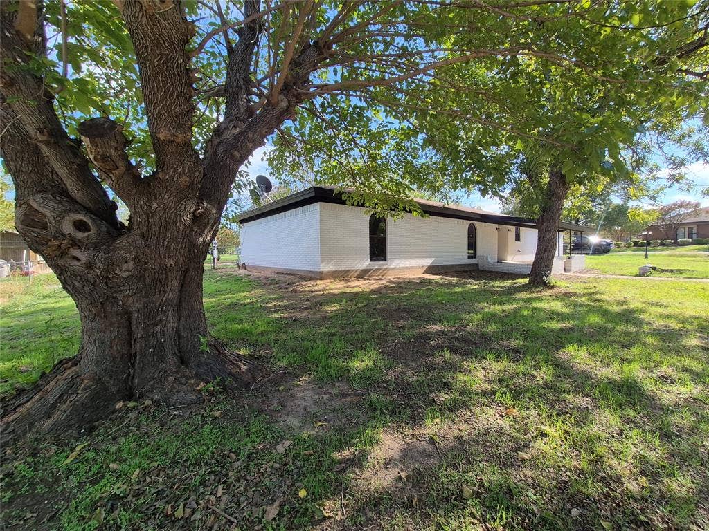 2029 Roberts Drive, Granbury, TX 76048 Single-story residence featuring white painted brick exterior, flat roofline, arched window detailing, and mature trees on a grass-covered lot