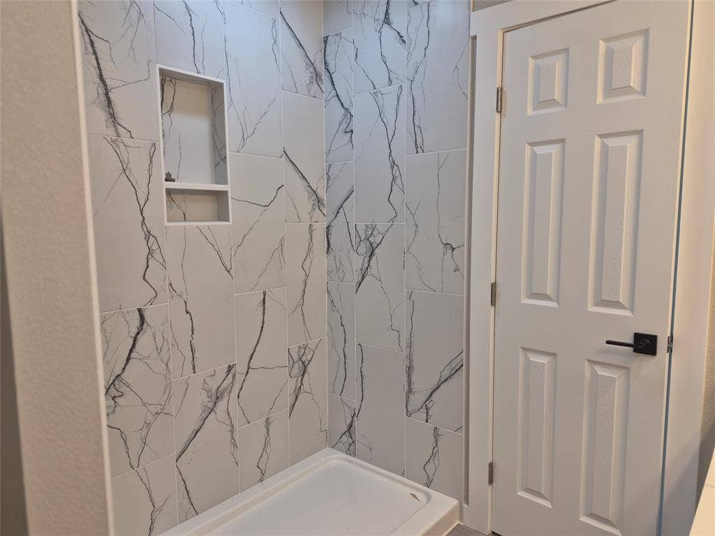 2029 Roberts Drive, Granbury, TX 76048 Shower enclosure featuring floor-to-ceiling large-format tile with decorative veining, built-in recessed storage niche, white acrylic shower basin, and a six-panel door with matte black hardware
