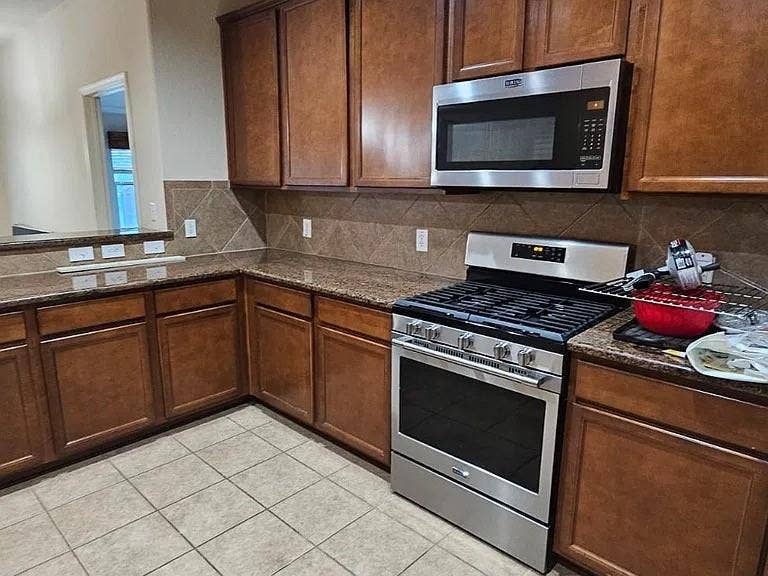 1000 Buffalo Creek Drive, McKinney, TX 75072 Kitchen featuring wood cabinetry, stone-finish countertops, ceramic tile backsplash, and stainless steel gas range with overhead microwave