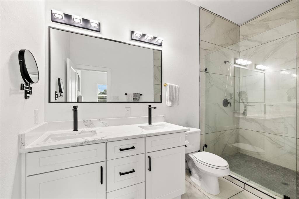 2502 Elsie Faye Heggins Street, Dallas, TX 75215 Dual-sink vanity with white cabinetry, stone-finish countertop, and matte black fixtures, paired with a glass-enclosed shower featuring large-format tile surround and matte black hardware