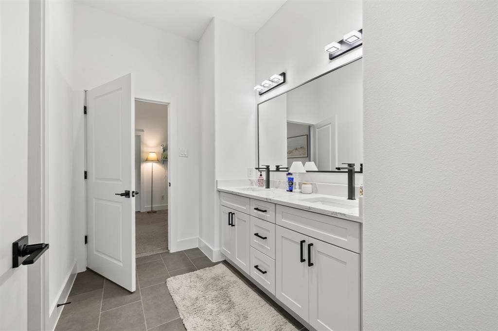 2502 Elsie Faye Heggins Street, Dallas, TX 75215 Dual-sink vanity featuring white cabinetry, stone-finish countertops, matte black hardware, and wall-mounted matte black faucets, complemented by two modern light fixtures and a full-width mirror