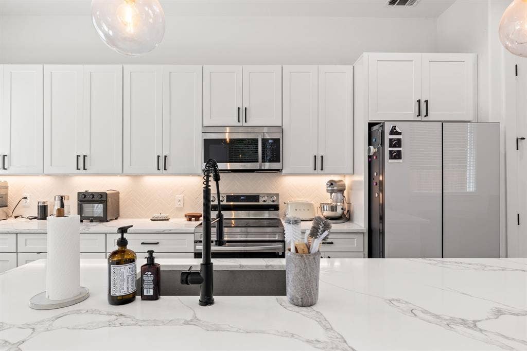 2502 Elsie Faye Heggins Street, Dallas, TX 75215 Kitchen featuring white Shaker-style cabinetry, a large island with a stone-surface countertop, herringbone-patterned backsplash, stainless steel appliances, and a matte black gooseneck faucet