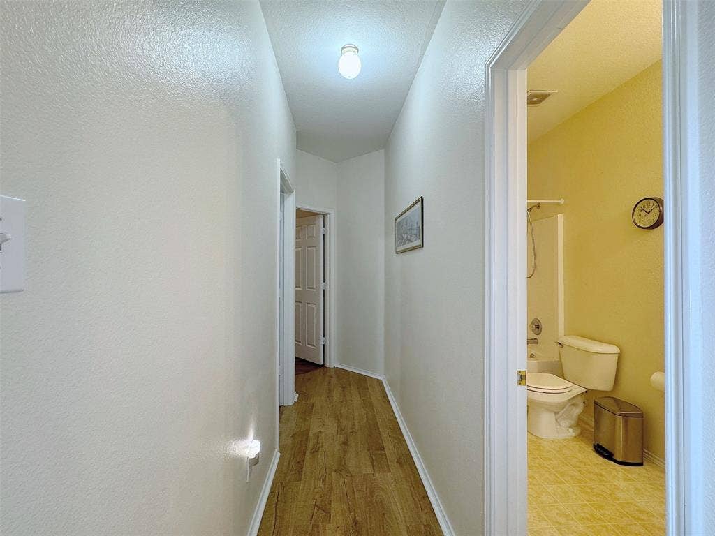 4102 Maidstone Drive, Garland, TX 75043 Interior hallway with wood-look flooring, ceiling lighting, and doorways connecting the adjoining rooms; a bathroom is visible at the side.