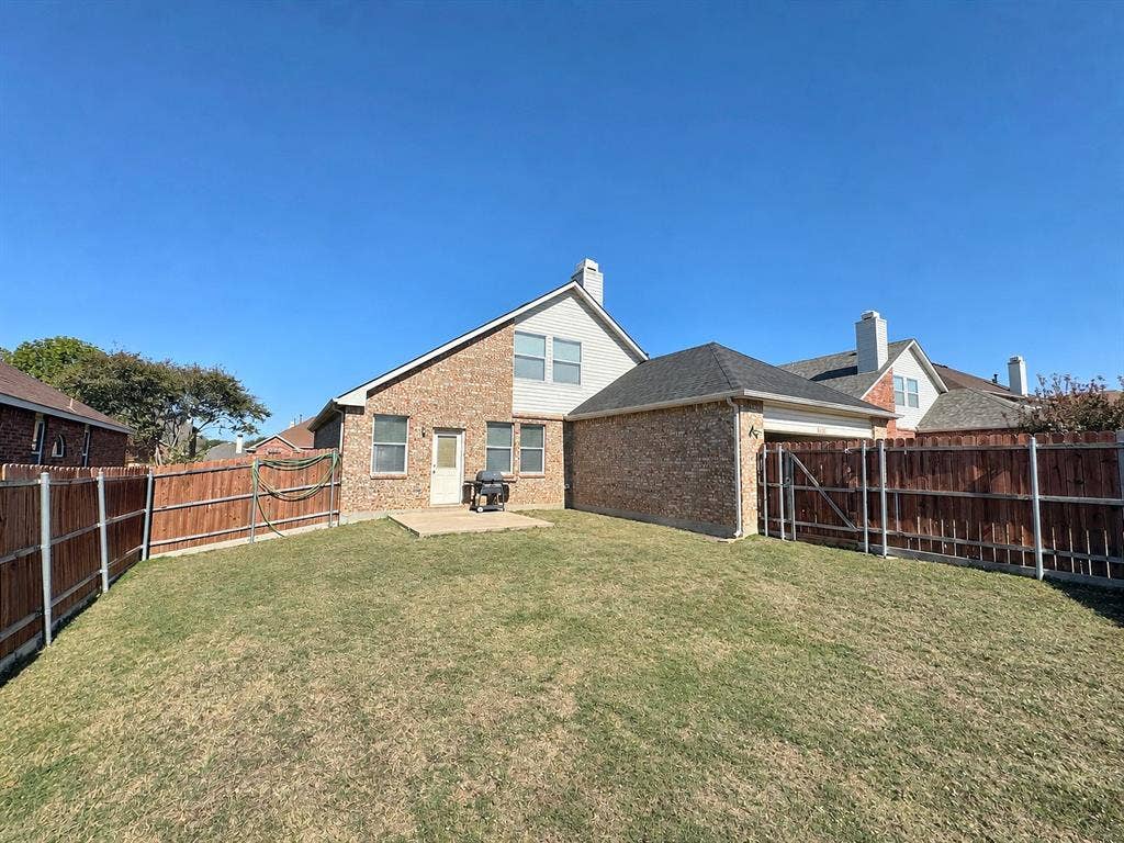 4102 Maidstone Drive, Garland, TX 75043 Ground-level backyard view showing the rear brick-and-siding exterior, lawn, patio area, and wood perimeter fencing.