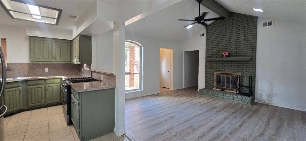 Kitchen featuring cabinetry, a gas cooktop, and stone countertops, paired with a living area showcasing wood-finish flooring, a vaulted ceiling, and a full-height brick fireplace