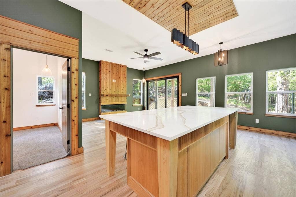 116 Carolynn Road, Mabank, TX 75156 Kitchen featuring an expansive island with a light-toned stone countertop, wood-finish flooring, a wood-paneled ceiling accent, a floor-to-ceiling wood fireplace surround, and multiple large windows
