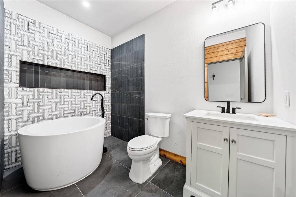 116 Carolynn Road, Mabank, TX 75156 Freestanding soaking tub with matte black wall-mounted faucet, geometric tiled accent wall featuring a recessed niche, white vanity with rectangular sink, and large-format dark floor tile