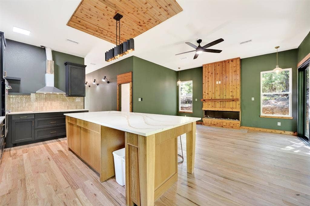 116 Carolynn Road, Mabank, TX 75156 Kitchen featuring an oversized island with a quartz countertop, wood-finish flooring, wood-paneled fireplace surround, integrated ceiling-mounted wood feature, and a modern linear pendant light fixture