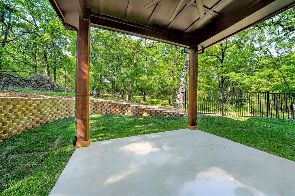 116 Carolynn Road, Mabank, TX 75156 Covered patio featuring concrete flooring and wooden support columns, complemented by a tiered stone retaining wall and black metal fencing