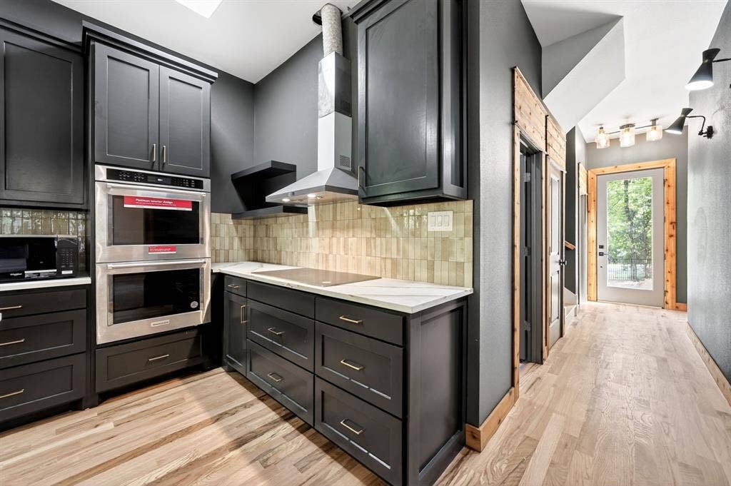 116 Carolynn Road, Mabank, TX 75156 Custom dark-finish cabinetry featuring brass-tone hardware, stainless steel built-in double ovens, natural stone-finish countertops, textured tile backsplash, and light wood-finish flooring