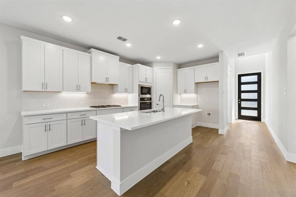 11512 Empress Drive, Frisco, TX 75035 Kitchen featuring white cabinetry, quartz countertops, wood-finish flooring, a herringbone-patterned tile backsplash, and a central island with a stainless steel sink