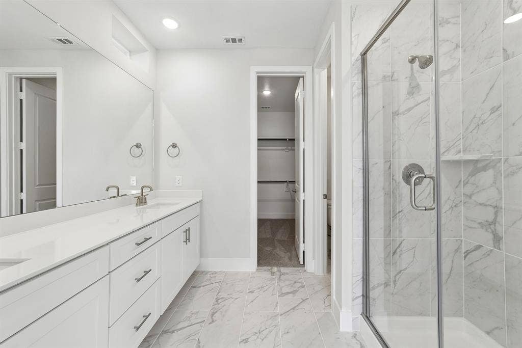 11512 Empress Drive, Frisco, TX 75035 Bathroom featuring white cabinetry, dual sinks with polished chrome fixtures, a full-width wall mirror, tile flooring with marble-pattern veining, and a glass-enclosed shower with tiled surround