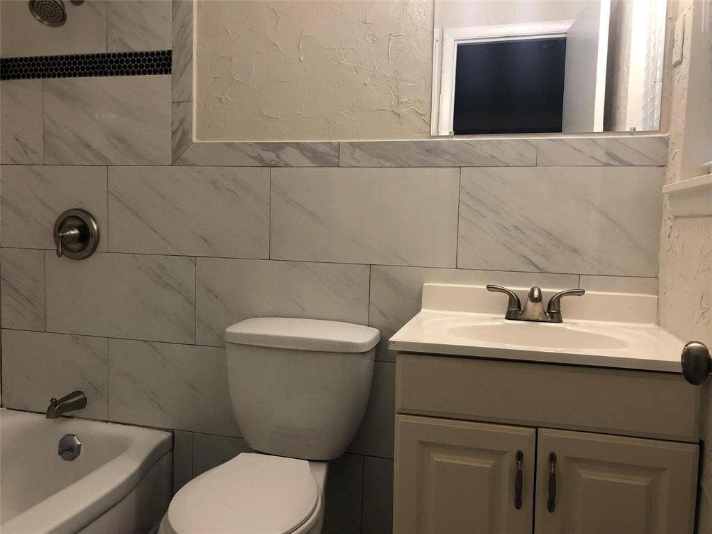 1217 Cloverdale Drive, Richardson, TX 75080 Bathroom featuring stone-finish wall tile, a built-in vanity cabinet with integrated sink, metallic double-handle faucet, and shower fixture with a decorative mosaic border