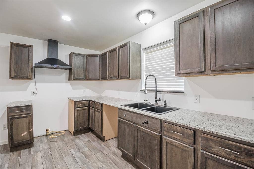 286 Larry Drive, Dallas, TX 75137 Kitchen featuring stained wood cabinetry, light-toned stone-finish countertops, dual-basin under-mount sink with a high-arch matte black faucet, wall-mounted range hood, and wood-finish flooring