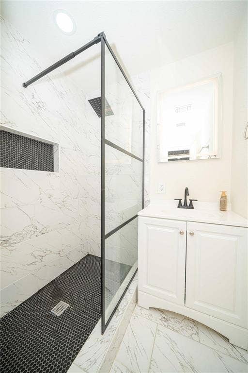 Walk-in shower with matte black-framed glass enclosure, stone-patterned wall tile, recessed tiled niche, and dark penny-round flooring, complemented by a white vanity with a top-mounted faucet and square mirror