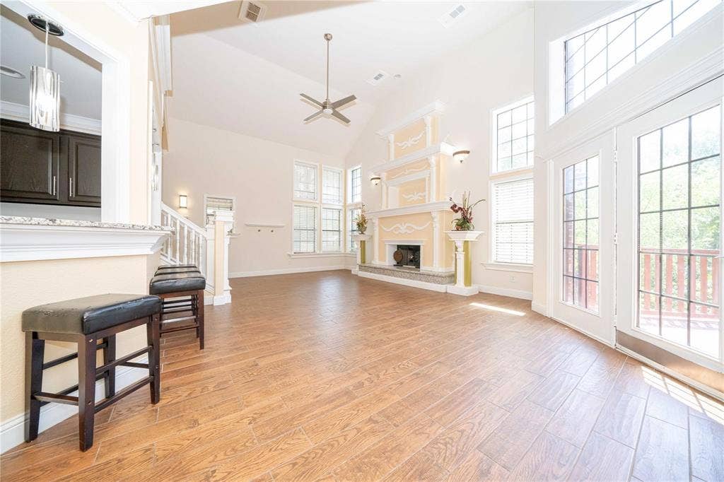 Open-concept living space featuring vaulted ceilings, wood-finish flooring, a decorative fireplace mantle with stone-surround hearth, and double glass-paneled exterior doors with an arched transom window