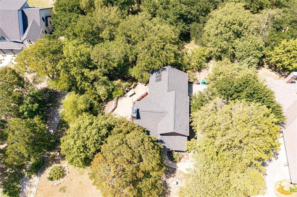 Aerial perspective featuring a multi-pitched asphalt shingle roof, solar panels, stone hardscaping, and mature trees on a wooded lot