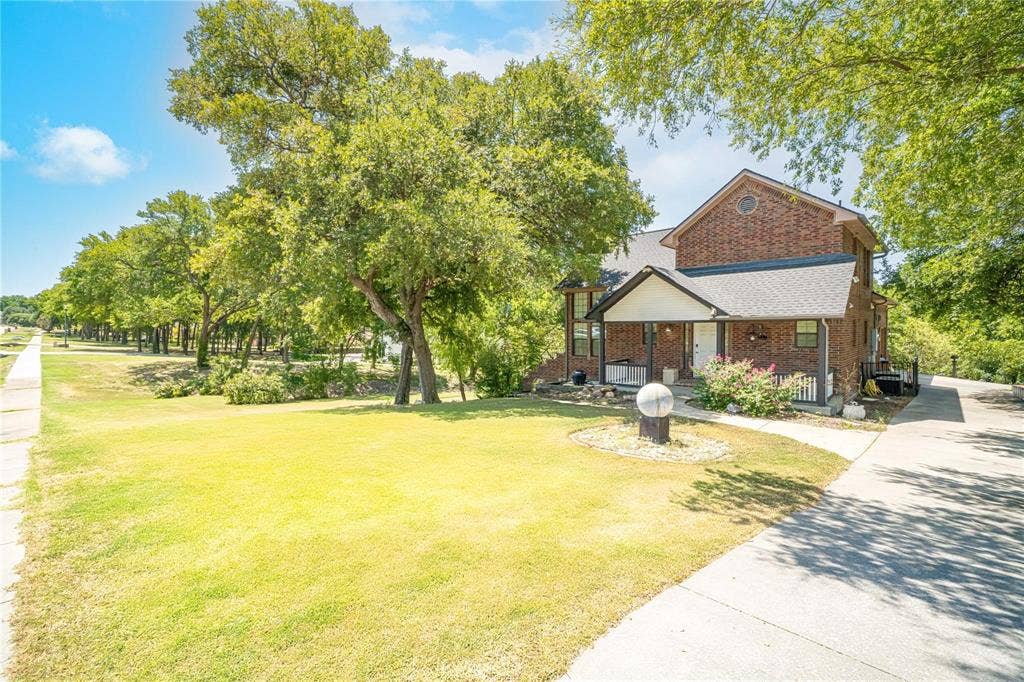 Two-story brick exterior featuring a covered front porch, concrete driveway, and expansive lawn with mature trees