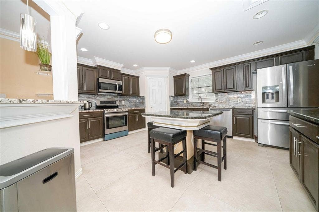 Kitchen featuring dark wood cabinetry, a central island with a stone countertop, stone-tiled backsplash, stainless steel appliances, and neutral tile flooring