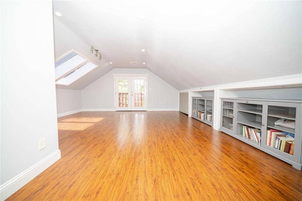Vaulted ceiling interior featuring wood-finish flooring, recessed lighting, double glass-panel doors, skylights, and built-in grey cabinetry