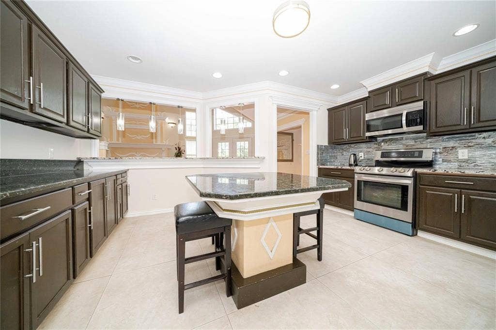 Kitchen featuring dark wood cabinetry, stone countertops, stainless steel appliances, a tiled backsplash, and a central island with custom millwork