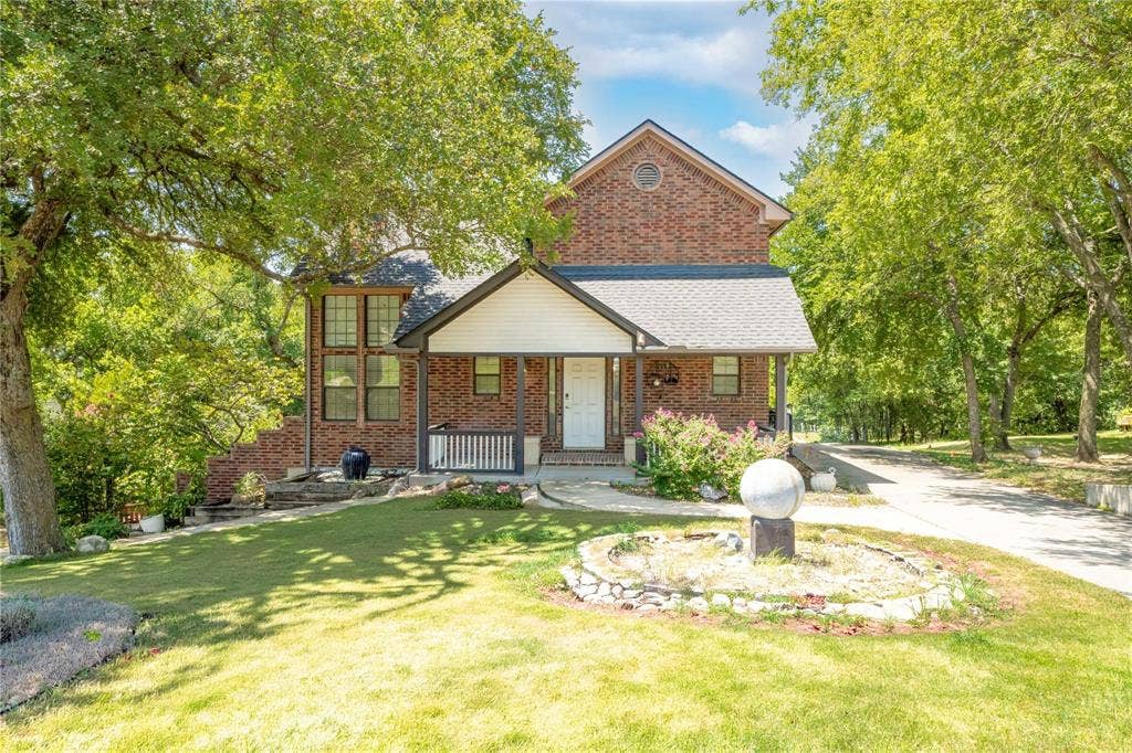 Multi-story brick exterior with a gabled roofline, covered front porch, stone-lined landscaping, and mature trees