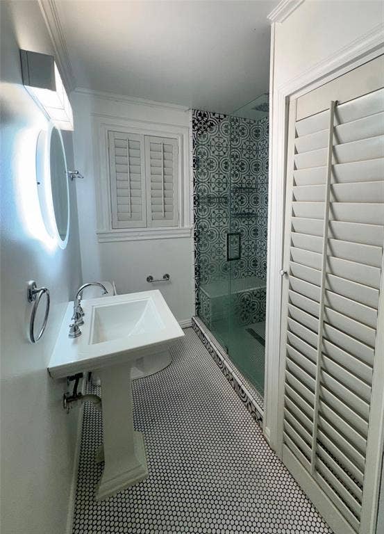 Pedestal sink with polished chrome faucet, glass-enclosed shower featuring patterned tile surround, plantation shutters, and monochromatic penny round floor tile