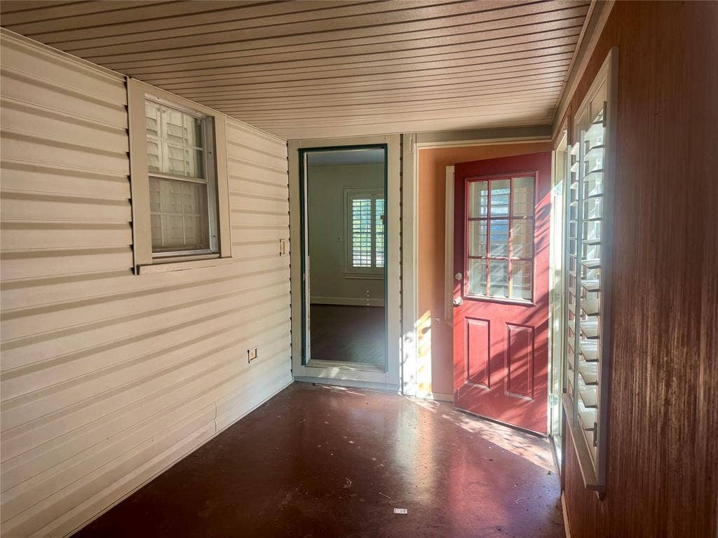 Covered porch featuring horizontal siding, wood-finish wall paneling, a red paneled entry door with glass light inserts, and windows with interior shutters