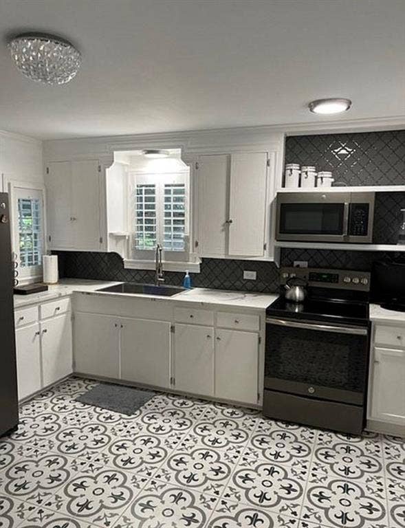 Kitchen featuring white cabinetry, patterned tile flooring, dark backsplash, stainless steel appliances, and interior window shutters