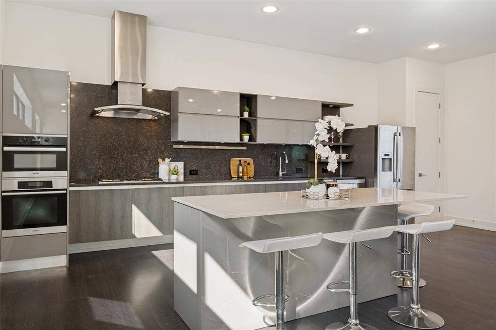 3336 Anchor Court, Dallas, TX 75219 Modern kitchen featuring flat-panel cabinetry, a central island with a quartz countertop, stainless steel appliances including a built-in double oven and range hood, and dark-toned wood-finish flooring