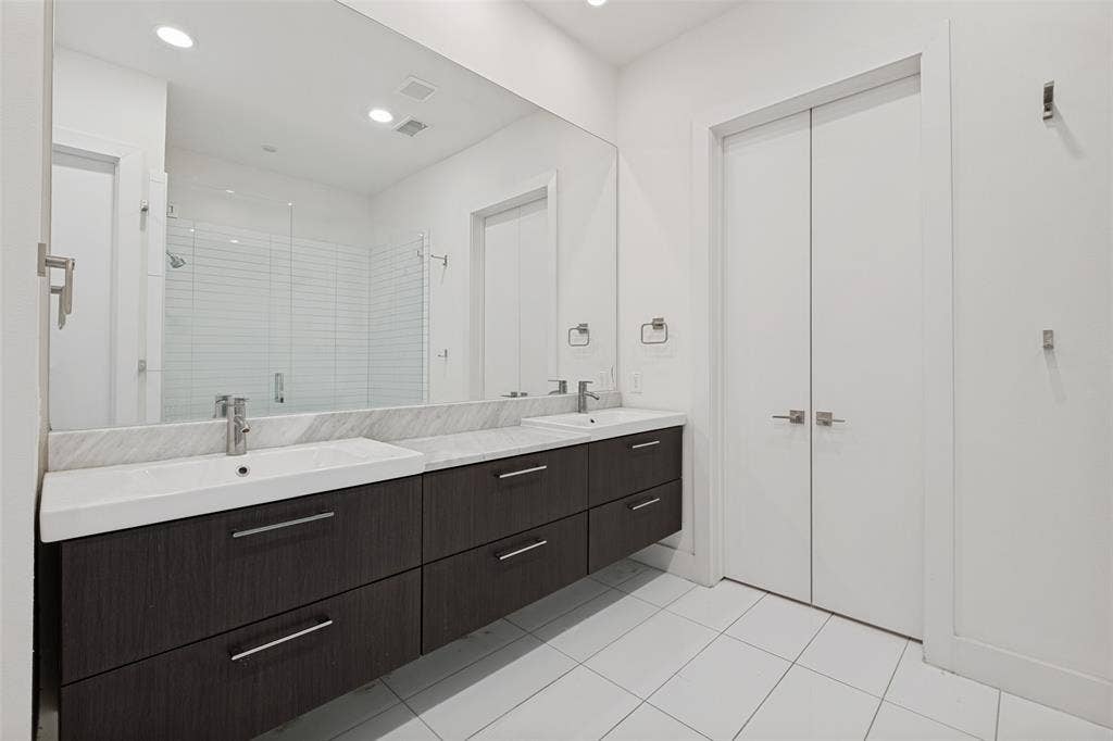 3336 Anchor Court, Dallas, TX 75219 Floating double vanity with dark wood-finish cabinetry, stone countertop, integrated sinks, and expansive wall-to-wall mirror, complemented by floor-to-ceiling white tile flooring