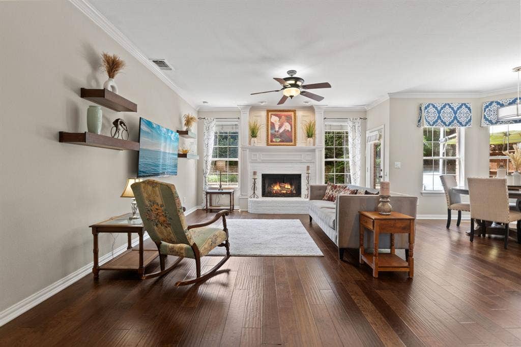 1110 Wedge Hill Road, McKinney, TX 75072 Open-concept living space featuring wood-finish flooring, a white-mantel fireplace with ornate columns, crown molding, a ceiling fan with integrated lighting, and multiple windows with deep sills