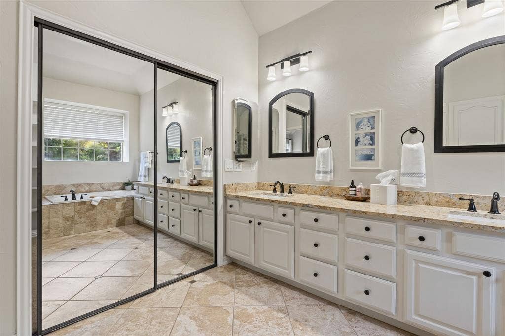 1110 Wedge Hill Road, McKinney, TX 75072 Dual vanity with stone-finish countertop, white shaker-style cabinetry, arched wall mirrors, stone tile flooring, and a jetted soaking tub with tile surround