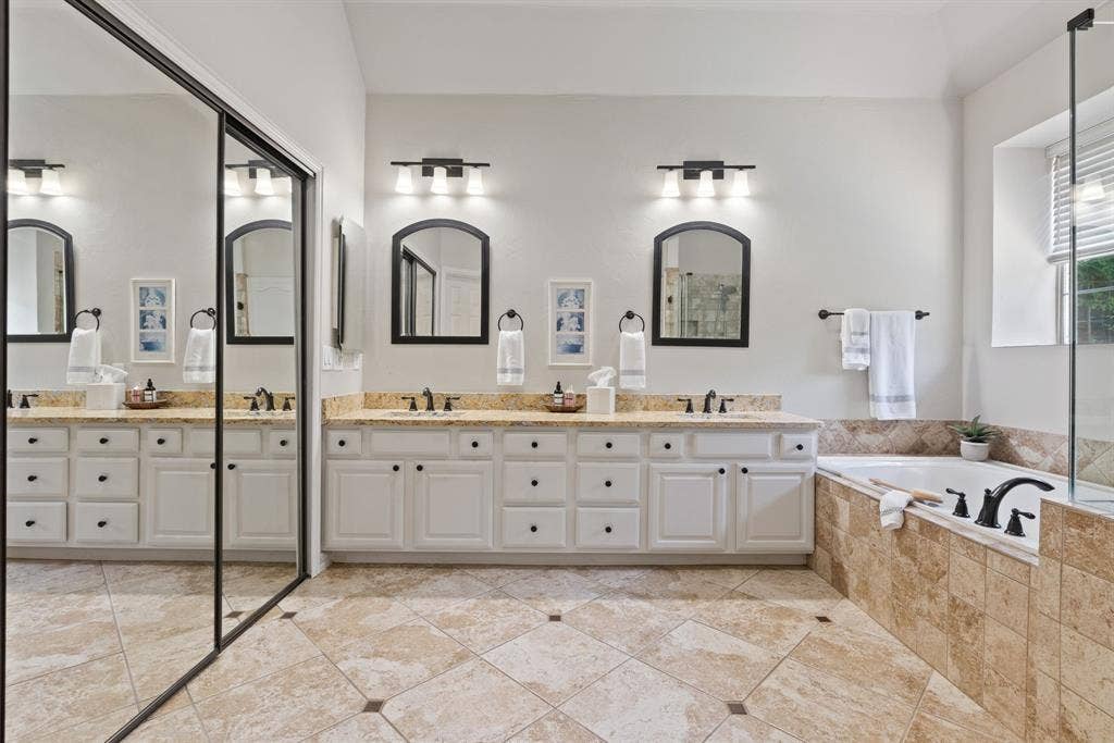 1110 Wedge Hill Road, McKinney, TX 75072 Dual-sink vanity with stone countertop, large-scale mirror wall, tile-surround soaking tub, and tile flooring with decorative inlay