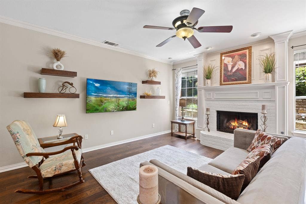 1110 Wedge Hill Road, McKinney, TX 75072 Living area featuring dark wood-finish flooring, a white brick fireplace with a custom mantel, built-in floating shelves, crown molding, and a ceiling fan