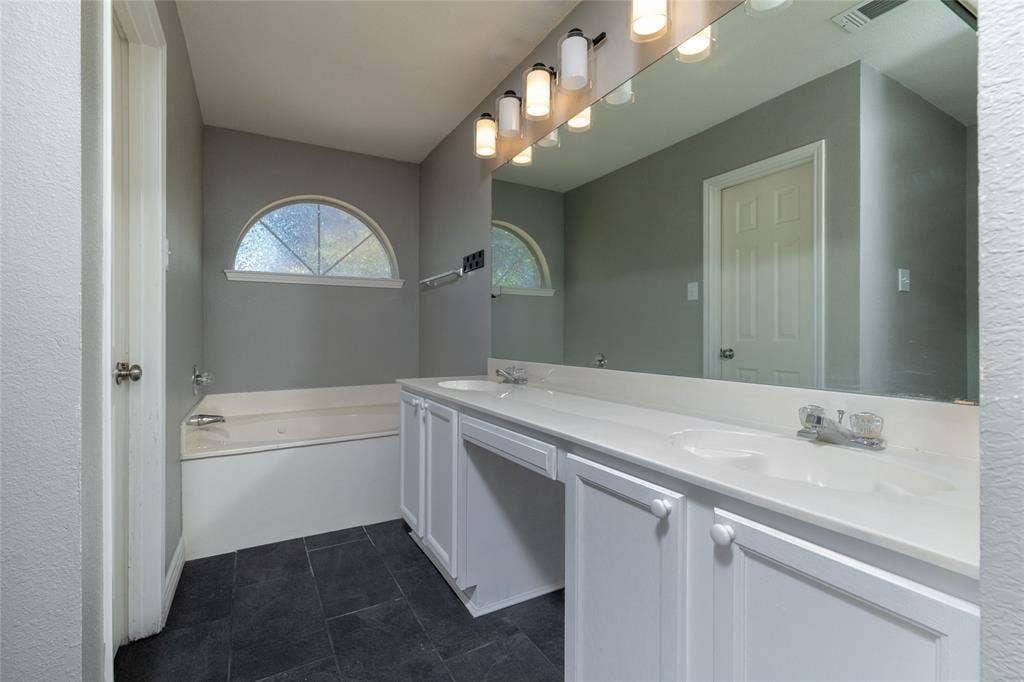 224 Cobblestone Circle, Red Oak, TX 75154 Dual-sink vanity with white cabinetry and a solid-surface countertop, full-width wall mirror, soaking tub, arched window, and stone-finish tile flooring