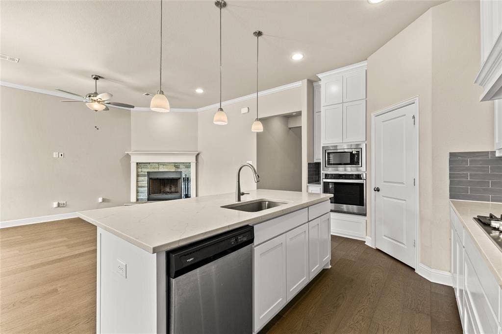 408 Abbott Lane, Waxahachie, TX 75165 Kitchen featuring a central island with a quartz countertop, white cabinetry, integrated stainless steel appliances, and wood-finish flooring, complemented by a stone-faced fireplace