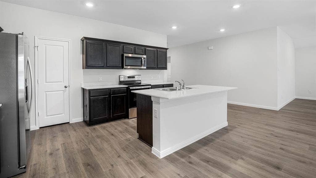 320 Ridan Drive, Waxahachie, TX 75165 Modern kitchen featuring dark wood cabinetry, stainless steel appliances, a white island with an integrated sink, light countertops, and wood-finish flooring