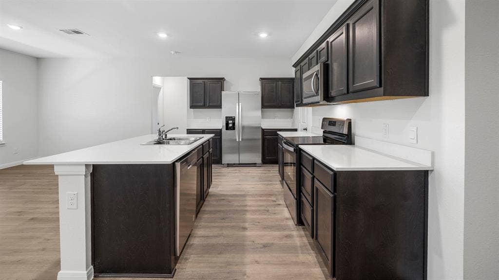 320 Ridan Drive, Waxahachie, TX 75165 Kitchen featuring an elongated island with a double basin sink, dark cabinetry, light-toned countertops, stainless steel appliances, and wood-finish flooring