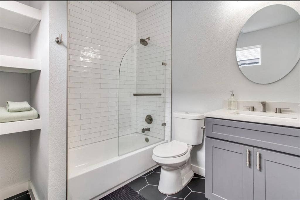 1606 W 12th Street, Dallas, TX 75208 Bathroom featuring a gray vanity with a stone countertop, white subway tile shower surround with an arched glass panel, built-in shelving, and black hexagonal floor tile