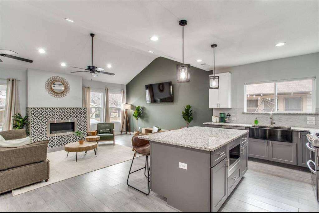 1606 W 12th Street, Dallas, TX 75208 Open-concept kitchen and living area featuring vaulted ceilings, wood-finish flooring, a patterned tile fireplace surround, an expansive center island with stone countertop, and charcoal-toned cabinetry