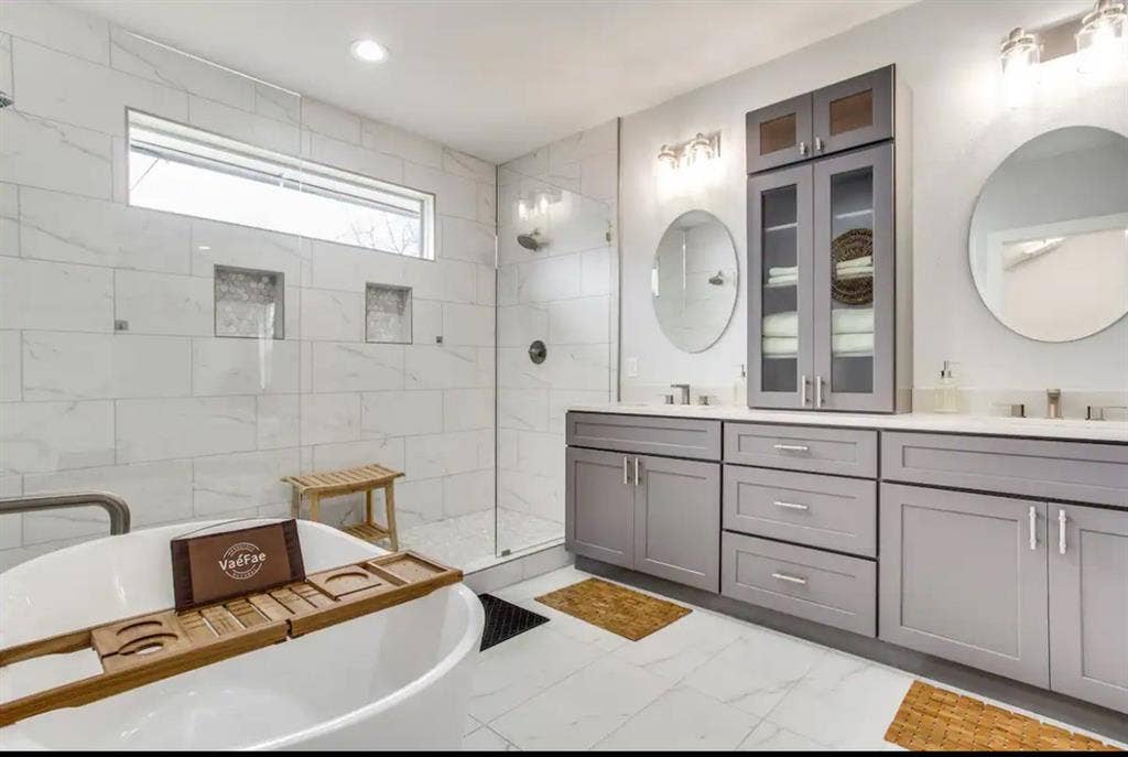 1606 W 12th Street, Dallas, TX 75208 Freestanding soaking tub, double-sink vanity with gray shaker-style cabinetry, glass-enclosed shower with tiled surround, built-in linen tower, and recessed shower niches