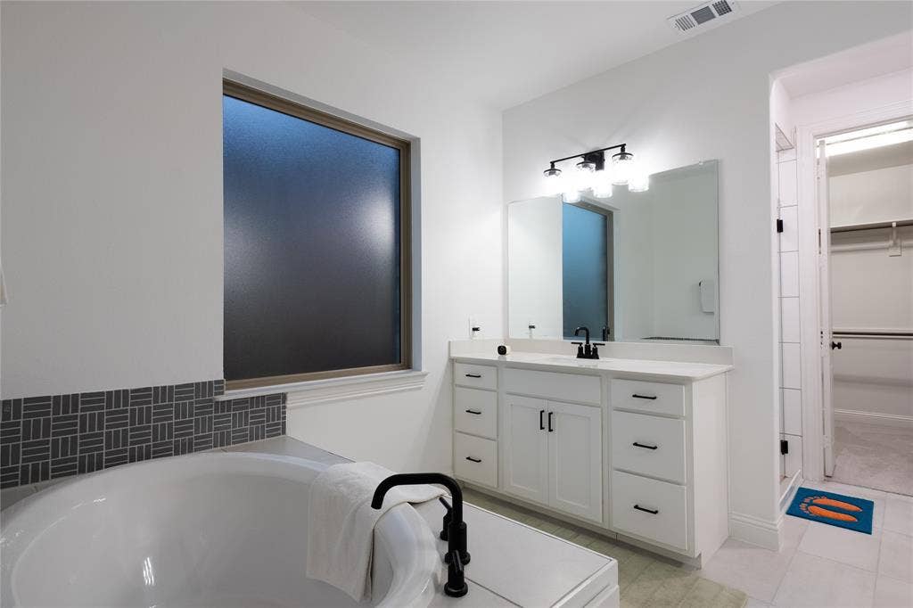 415 Rock Rose Lane, Wylie, TX 75098 Built-in white vanity with stone-style countertop, undermount sink with matte black hardware, frosted window, deep soaking tub with matte black faucet, and patterned decorative tile backsplash