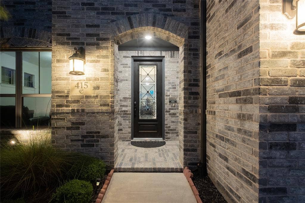 415 Rock Rose Lane, Wylie, TX 75098 Exterior entryway featuring a multi-tonal brick facade, an arched masonry header, a dark-finished front door with decorative glass inlay, and wall-mounted exterior sconces