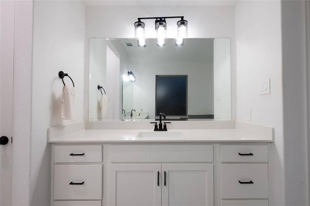 415 Rock Rose Lane, Wylie, TX 75098 Bathroom vanity featuring white cabinetry, a solid-surface countertop with an integrated basin, a large plate glass mirror, matte black hardware, and a three-light fixture with textured glass shades