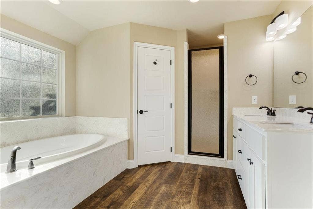 401 Mesa, Lone Oak, TX 75453 Bathroom featuring a deep soaking tub with a stone-finish surround, dual-sink vanity with light-toned cabinetry, wood-finish flooring, glass-enclosed shower, and multi-light wall sconce
