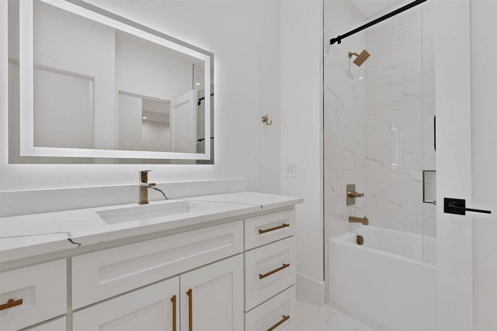 7503 Robin Road, Dallas, TX 75209 Shaker-style white vanity with a quartz countertop, integrated LED-backlit mirror, brass-finish hardware, and a tub-shower combination featuring tile surround and a frameless glass door