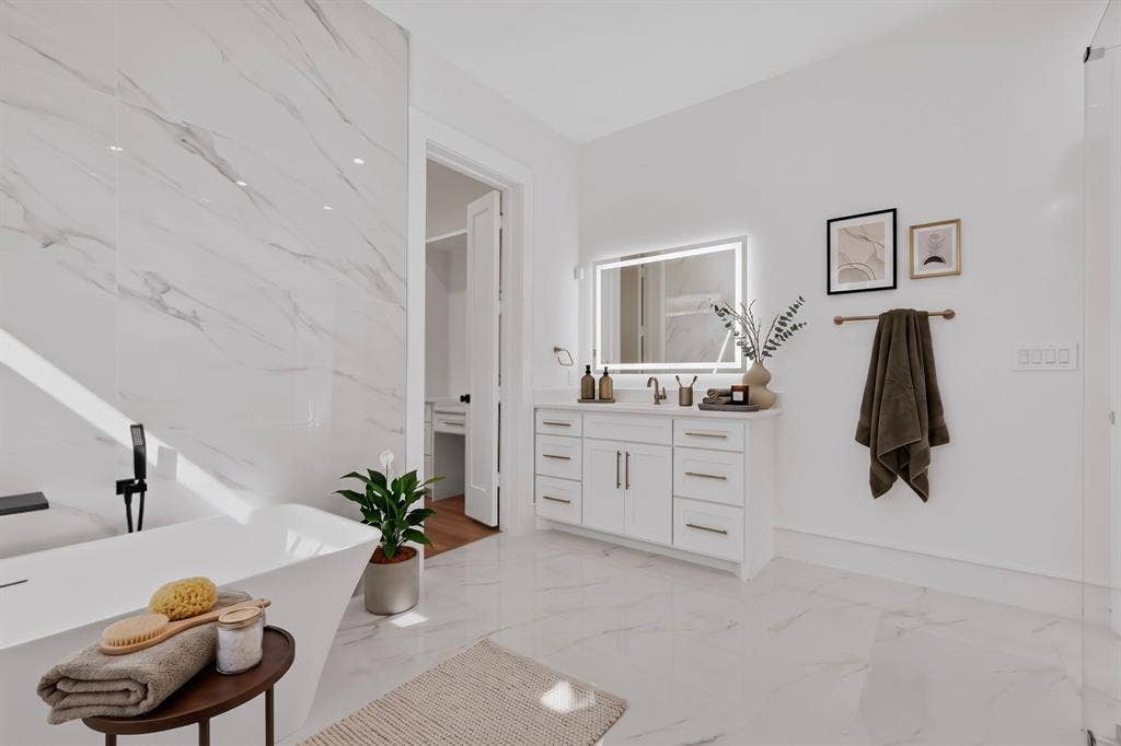 7503 Robin Road, Dallas, TX 75209 Freestanding soaking tub with a wall-mounted faucet, large-format stone-finish tile flooring, white shaker-style vanity with metal hardware, and an illuminated wall-mounted mirror