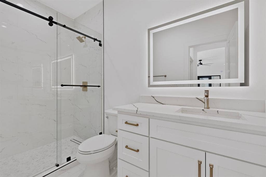 7503 Robin Road, Dallas, TX 75209 Walk-in shower with floor-to-ceiling veined tile, frameless glass sliding enclosure, brass-finish fixtures, quartz vanity countertop, and backlit wall-mounted mirror